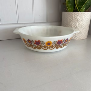 Può includere: Ciotola Pyrex bianca di forma ovale con un motivo floreale arancione, viola e giallo. La ciotola ha due manici e un bordo dorato. Una pianta decorativa in un vaso bianco è sullo sfondo.