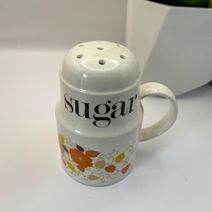 Op de afbeelding: Een witte keramische suikerdispenser met een bloemenpatroon in oranje, geel en bruin. De dispenser heeft een handvat en staat "sugar" in zwarte letters.