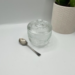 May include: A clear glass jar with a lid, featuring a textured design. The jar is sitting on a white surface, and a silver spoon is lying next to it.