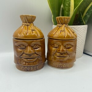 Puede incluir: Dos botes de cerámica marrón con caras, uno con la etiqueta "COFFEE" y el otro "SUGAR". Las tapas tienen forma de sacos atados. Los botes tienen un estilo vintage y de arte popular.