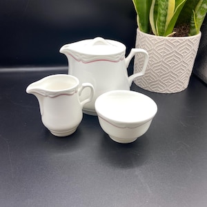 Könnte beinhalten: Weißes Keramik-Set aus Teekanne, Milchkännchen und Zuckerdose mit einem dünnen roten Streifen am Rand jedes Teils.