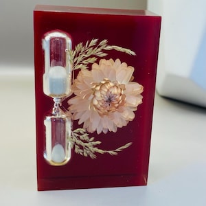 Puede incluir: Un reloj de arena decorativo encerrado en un bloque rectangular de resina rojo oscuro. El reloj de arena es plateado y contiene arena blanca. Una flor seca y ramitas están incrustadas en la resina, agregando un elemento natural. Altura: 7 cm.