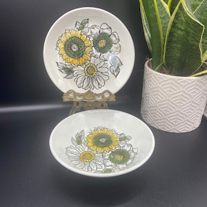 Könnte beinhalten: Zwei weiße Keramikschalen mit einem Blumendesign mit gelben Sonnenblumen, grünen und weißen Blumen. Eine Schale steht auf einem dekorativen Ständer. Die Schalen sind Teil eines Küchengeschirrsets.