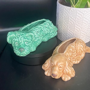 Könnte beinhalten: Zwei keramische Pflanzgefäße in Hundeform. Eines ist hellgrün, das andere ist hellbraun. Beide Pflanzgefäße haben eine lange, rechteckige Öffnung für Pflanzen. Die Pflanzgefäße stehen auf einer schwarzen Oberfläche, mit einem weißen Topf mit Muster und einer Pflanze im Hintergrund.