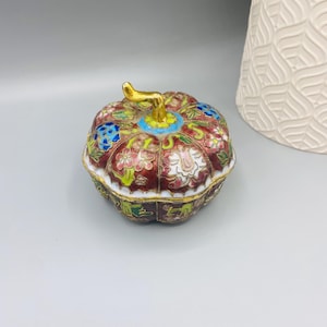 Puede incluir: Una caja de baratijas decorativa con forma de calabaza y un tallo dorado. La caja presenta un fondo granate con motivos florales en blanco, azul y rosa. El borde es blanco y la base tiene detalles verdes y dorados. Una pieza única y ornamentada.