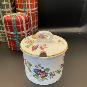 Puede incluir: Un azucarero de porcelana blanca con tapa y diseño floral. La tapa tiene una pequeña asa y un borde dorado. El tazón tiene un borde dorado alrededor de la parte superior y un diseño floral con pájaros.