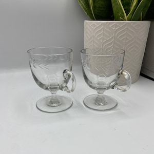 Puede incluir: Dos tazas de vidrio transparente con diseños florales grabados y asas. Las tazas están sobre una superficie blanca.