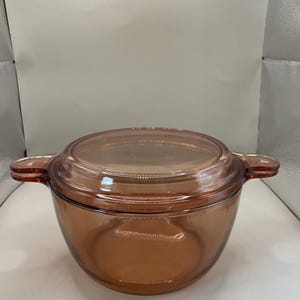 Puede incluir: Una fuente de horno de vidrio transparente de color ámbar con una tapa a juego. La fuente tiene dos asas laterales y una forma redondeada. La tapa encaja a ras. Esta fuente es adecuada para hornear y servir.