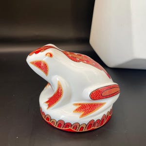 May include: A white ceramic frog ornament with red and gold patterns. The frog features detailed designs on its back, legs, and base. This decorative figurine is likely for display.