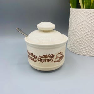 May include: Cream-colored ceramic pickle and chutney jar with a lid and a small spoon. The jar has a brown graphic and text that reads "Pickles & Chutney". The jar is next to a white decorative vase.