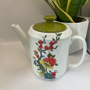 May include: White ceramic teapot with a green lid and floral design. The teapot features a floral pattern with red, yellow, blue, and green flowers.