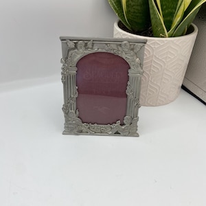 Puede incluir: Un marco de fotos plateado con un diseño ornamentado con querubines y acentos florales. El marco tiene una forma rectangular y una alfombra de terciopelo rojo.