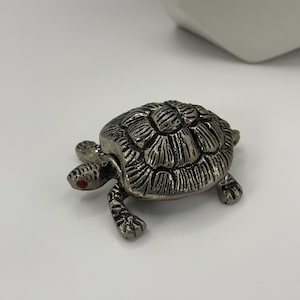 May include: A silver turtle figurine with a detailed shell and red eyes. The turtle is sitting on a white surface.
