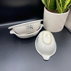 May include: Three white ceramic bowls with handles, featuring a black graphic and text. The bowls are oval-shaped with a small spout on one end. A white patterned pot with a green plant is in the background.