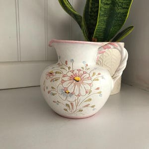 Könnte beinhalten: Eine weiße Keramikkanne mit rosafarbenem Rand und Blumenmuster. Die Kanne hat einen Henkel und ist mit rosa, blauen und gelben Blumen verziert.