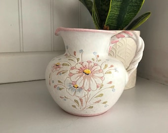 May include: A white ceramic pitcher with a pink rim and floral design. The pitcher has a handle and is decorated with pink, blue, and yellow flowers.