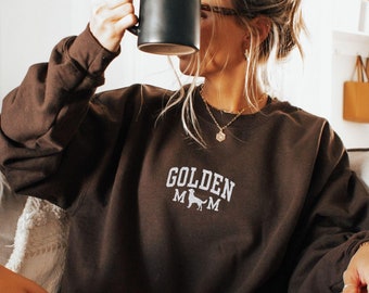May include: A woman wearing a brown sweatshirt with the text "Golden Mom" and a dog silhouette. She is sitting on a couch and holding a black mug.