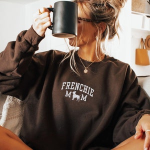 May include: A woman wearing a brown sweatshirt with the text "Frenchie Mom" and a small dog silhouette. She is sitting on a couch and holding a black mug.