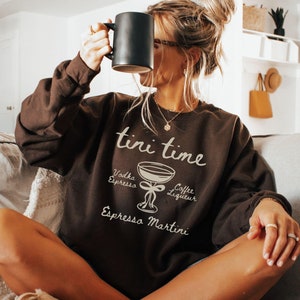 May include: A brown sweatshirt with the text "tini time" and a graphic of a martini glass with the ingredients listed: Vodka, Espresso, Coffee Liqueur, Espresso Martini.