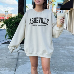 Pode incluir: Uma camisa de moletom cor creme com o texto "Asheville North Carolina" estampado na frente. A camisa de moletom est&aacute; sendo usada por uma pessoa com t&ecirc;nis branco.