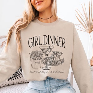 May include: A beige sweatshirt with the text "GIRL DINNER" and a graphic of a martini glass, pizza, and fries. The text below the graphic reads "It's a Good Day for a Girl Dinner."