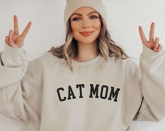 May include: A woman wearing a beige sweatshirt with the text "CAT MOM" in black lettering. She is wearing a beige beanie and has her hands up in a peace sign.