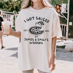 May include: A white oversized t-shirt with black text that reads "I got Sauced est 2025 at James & Emily's Wedding". The shirt features a black and white illustration of a pizza slice.