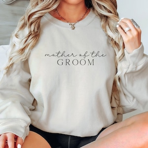 May include: A light beige sweatshirt with the text "mother of the groom" in black script.