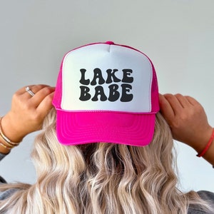 May include: Pink trucker hat with a white mesh back and black retro font that says "LAKE BABE".