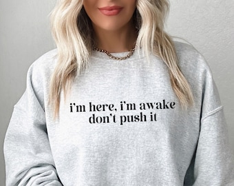 May include: A light gray sweatshirt with the text "I'm here, I'm awake don't push it" in black. The sweatshirt has long sleeves and a crew neck. The model is wearing a gold chain necklace.