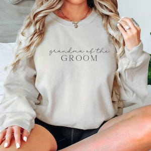 May include: A light beige sweatshirt with the text "grandma of the groom" printed in black.