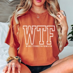 Op de afbeelding: Een roestkleurig T-shirt met de tekst "WTF" in witte omrande letters. De tekst eronder luidt "WINE TURKEY FAMILY" met hartjes tussen de woorden.