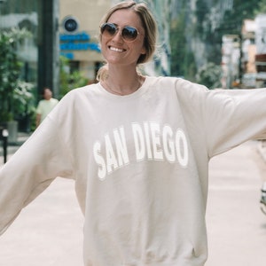 Pode incluir: Uma camisa de moletom de cor creme com o texto "SAN DIEGO" impresso em branco na frente.