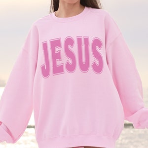 May include: Pink sweatshirt with the word "JESUS" printed in a bold, pink font.