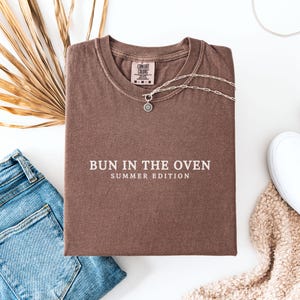 May include: A brown t-shirt with the text "Bun in the Oven Summer Edition" printed in white.