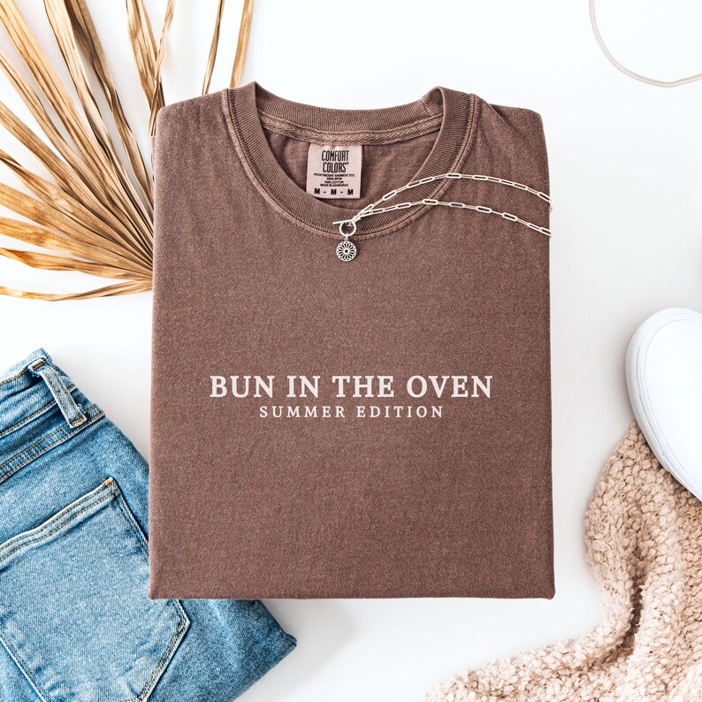 May include: A brown t-shirt with the text "Bun in the Oven Summer Edition" printed in white.