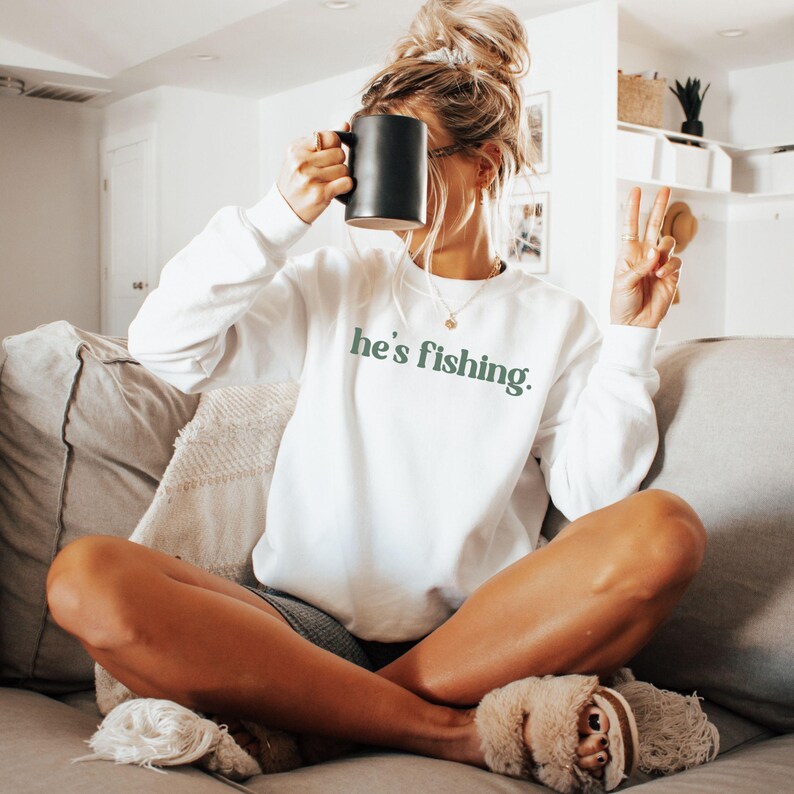 Puede incluir: Una sudadera blanca con el texto "he's fishing" en letras verdes. La modelo lleva la sudadera y sostiene una taza negra.