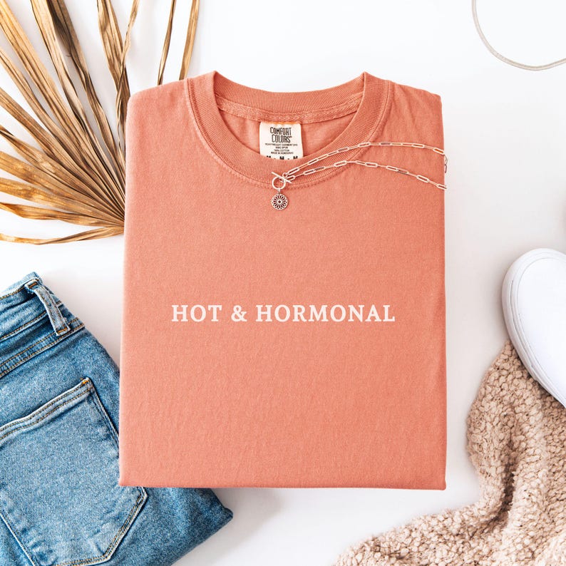 May include: A coral colored t-shirt with the text "HOT & HORMONAL" printed in white on the front. The shirt is folded and laid flat on a white surface.