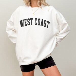 Puede incluir: Sudadera blanca de gran tama&ntilde;o con texto negro que dice "WEST COAST" en una fuente universitaria.