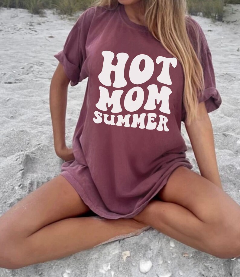 May include: A maroon t-shirt with the text "HOT MOM SUMMER" in white, retro-style font. The shirt is worn by a person sitting on a sandy beach.