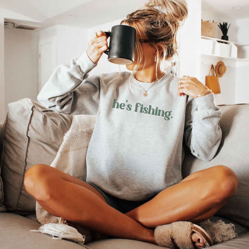 Puede incluir: Una sudadera gris con el texto "he's fishing" en letras verdes. La sudadera la lleva puesta una persona sentada en un sof&aacute;.