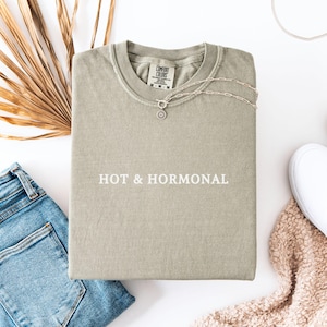 May include: A light green t-shirt with the text "HOT & HORMONAL" printed in white on the front. The shirt is laid flat on a white surface.