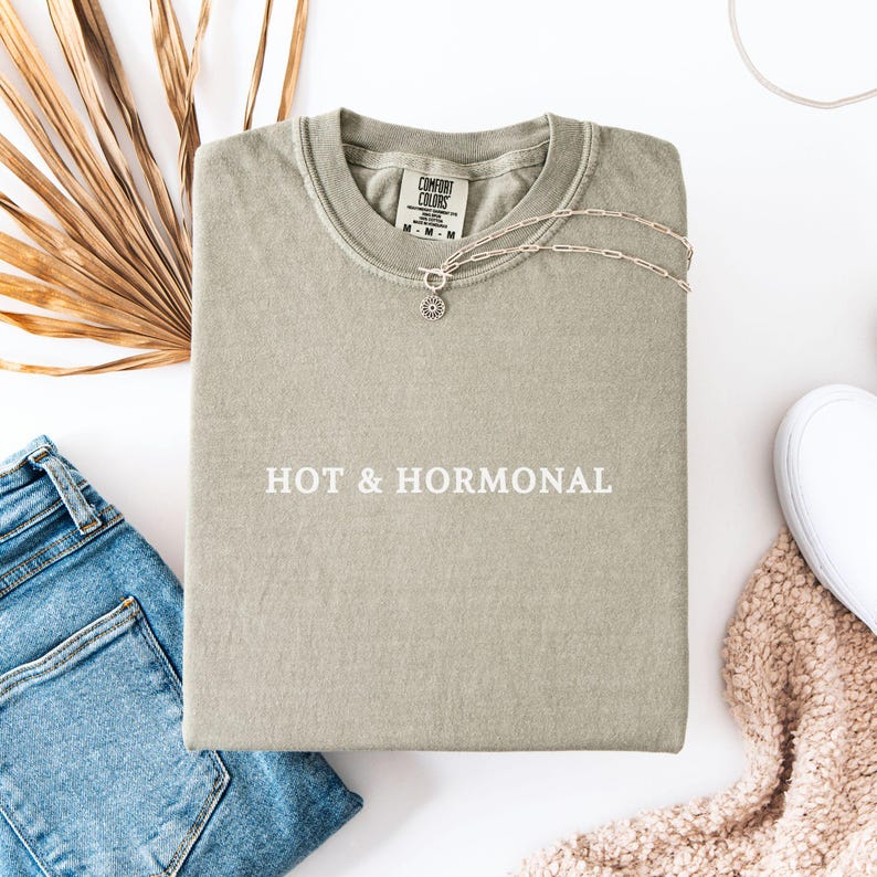 May include: A light green t-shirt with the text "HOT & HORMONAL" printed in white on the front. The shirt is laid flat on a white surface.
