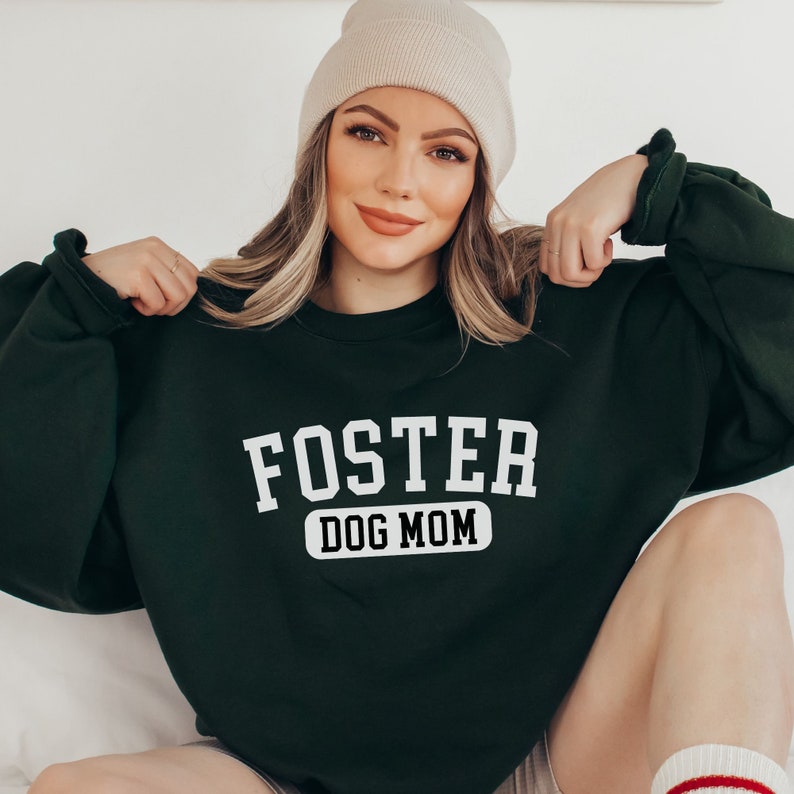 May include: A dark green sweatshirt with the text "FOSTER DOG MOM" in white lettering. The sweatshirt has a relaxed fit and a crew neck.