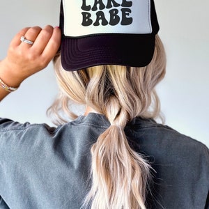 May include: A black and white trucker hat with the text "LAKE BABE" in a retro font.