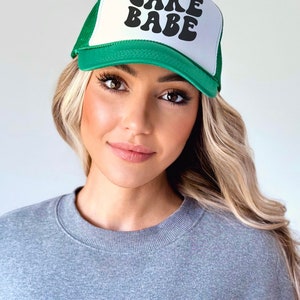 May include: A green and white trucker hat with the text "LAKE BABE" in black retro-style lettering.