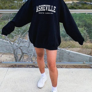 Pode incluir: Uma camisa de moletom preta com texto branco que diz "Asheville, Carolina do Norte". A modelo est&aacute; usando a camisa de moletom com t&ecirc;nis brancos.