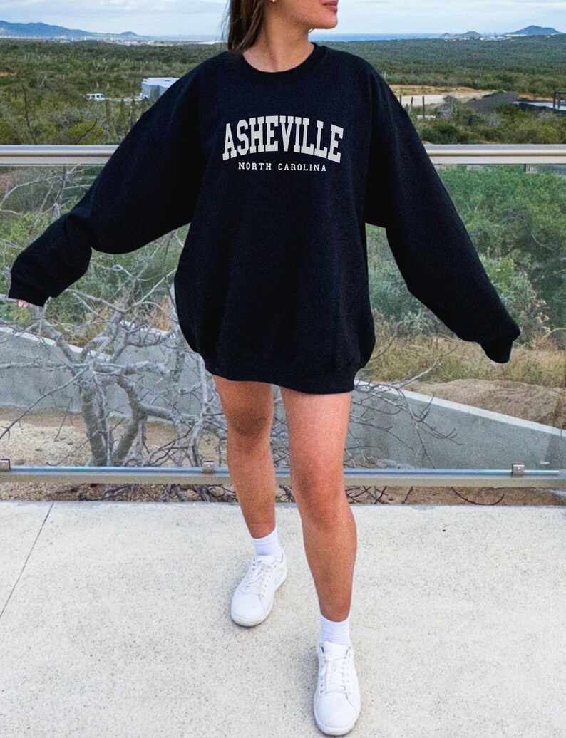 Pode incluir: Uma camisa de moletom preta com texto branco que diz "Asheville, Carolina do Norte". A modelo est&aacute; usando a camisa de moletom com t&ecirc;nis brancos.