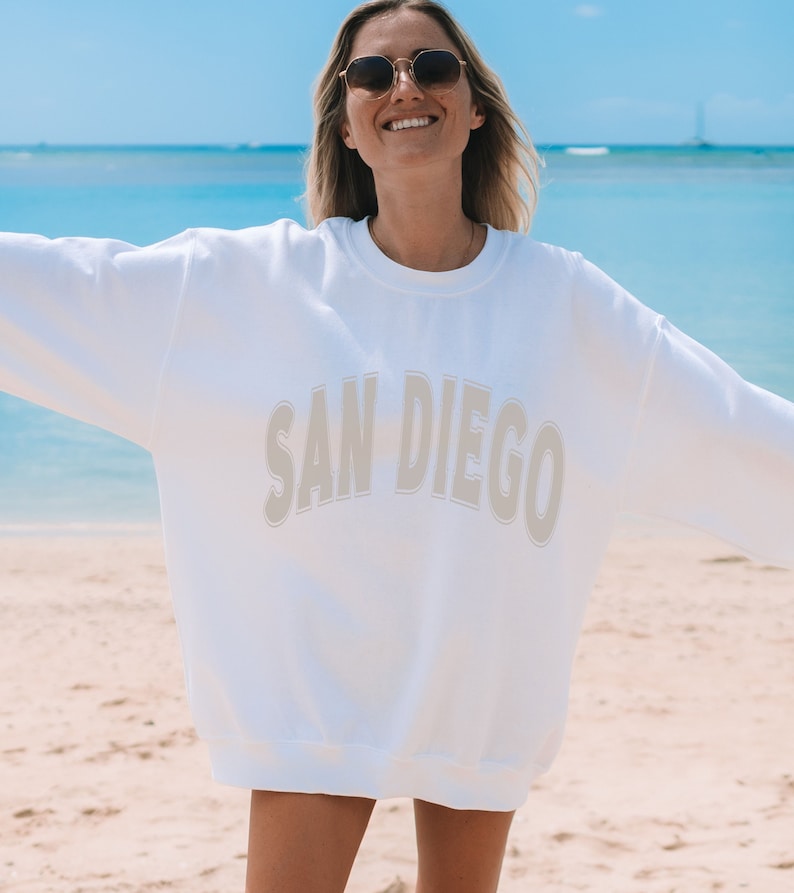 May include: A white sweatshirt with the text "SAN DIEGO" in a beige color on the front. The sweatshirt is worn by a person standing on a beach.