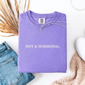 May include: A purple t-shirt with the text "HOT & HORMONAL" printed in white on the front.